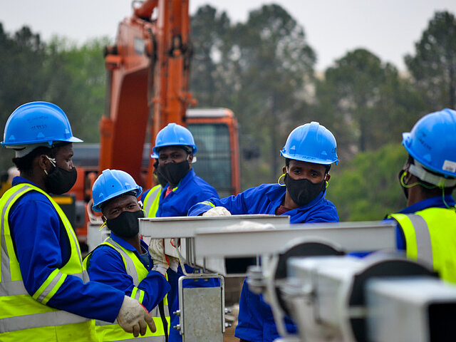 A group of workers is assembling the stands on which the solar panels will later be mounted. In the background, there is a construction machine.